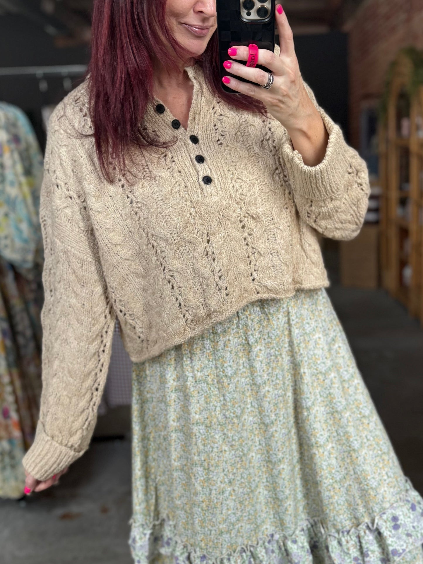 A woman takes a mirror selfie in a clothing boutique, wearing a beige cable-knit cropped cardigan over a light sage green and white floral tiered maxi skirt with ruffle trim, paired with beige pointed-toe heels.